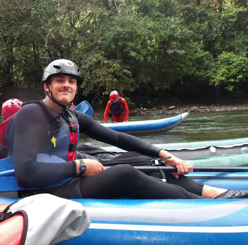 A man wearing a helmet and wetsuit is sitting in a blue kayak on a river. He is holding a paddle and looking at the camera with a slight smile. In the background, another person in a red wetsuit is in a kayak. The river is surrounded by lush green trees.